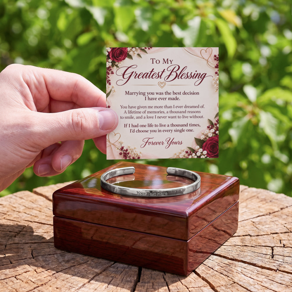 A hand holds a printed remembrance card above a polished wooden box containing a silver engraved cuff bracelet, set on a tree stump with a blurred green outdoor background.