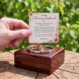 A hand holds a floral sympathy card above a polished wooden box containing a silver engraved cuff bracelet, set on a rustic tree stump with a blurred green garden background.