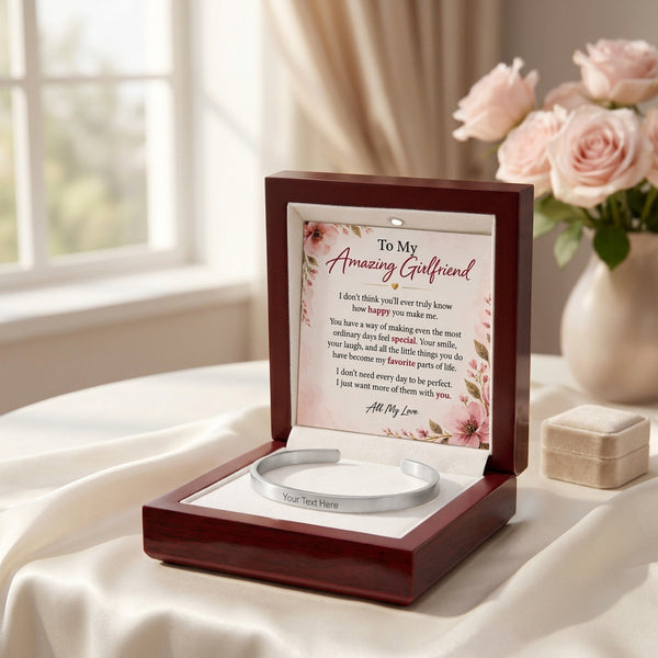 A silver engraved bracelet presented in an open dark wood box with a floral message card inside, styled on draped white fabric beside pink roses and a small matching box.