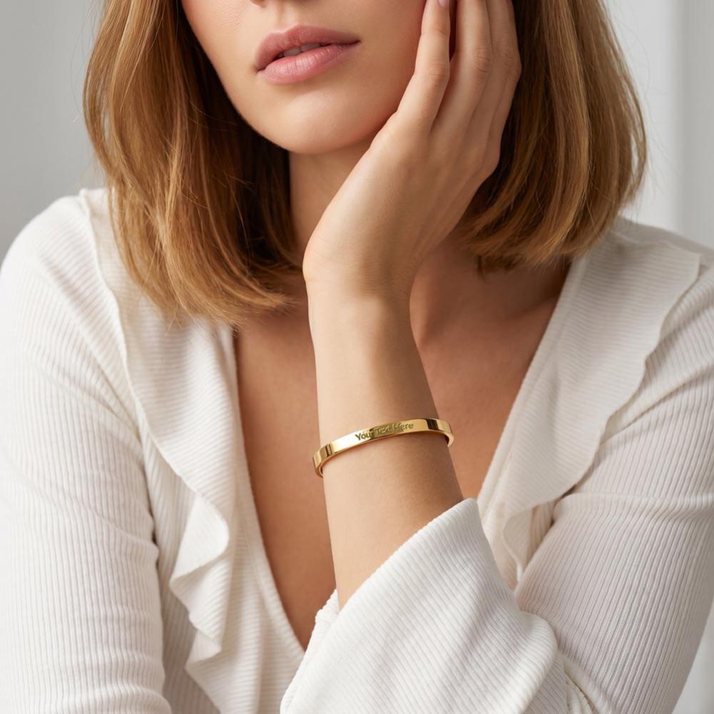 A close-up of a woman wearing a slim gold bangle bracelet with engraved lettering, paired with a white ribbed blouse.