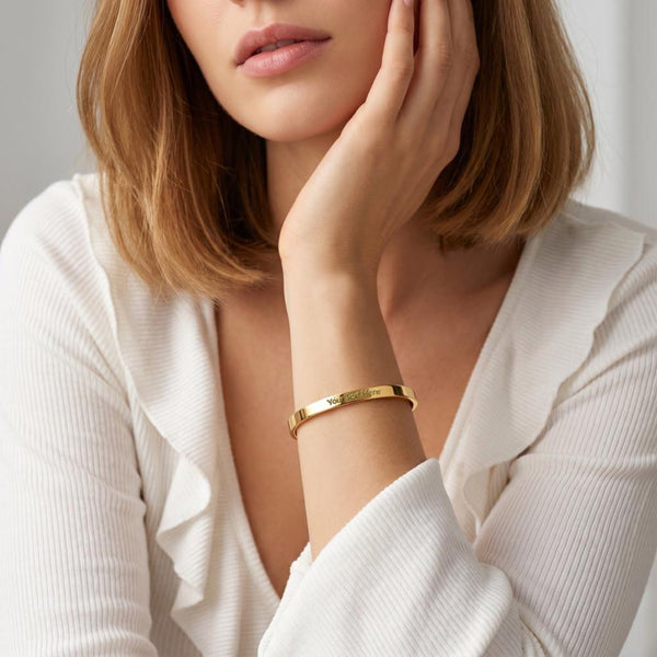 A close-up of a woman wearing a slim gold bangle bracelet with engraved lettering, paired with a white ribbed blouse.