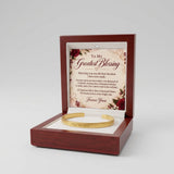 A gold-colored open cuff bracelet gift box with a reddish-brown wooden frame, featuring a sentimental 'To My Greatest Blessing' message card and a white interior cushion holding the bracelet.