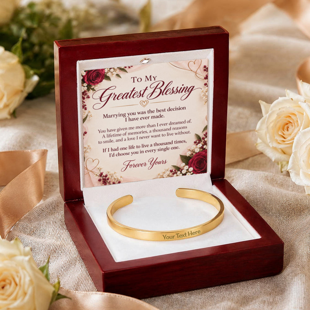A gold cuff bracelet displayed inside an open dark red wooden gift box with a floral message card. The card reads 'To My Greatest Blessing' and includes a romantic message, with the bracelet resting on a white insert.