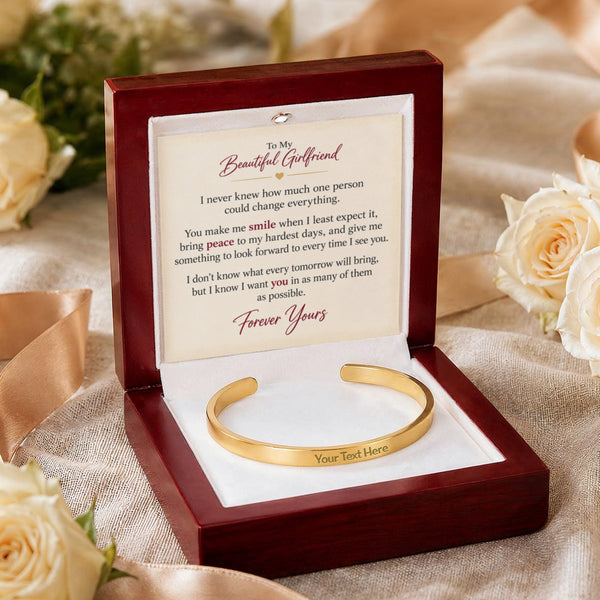A gold cuff bracelet presented in an open dark wood gift box with a white insert and a printed message card addressed to a beautiful girlfriend. The box is styled on draped satin fabric with cream roses in the background.
