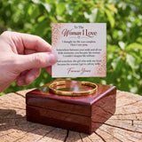 A hand holds a printed sentimental card above a polished wooden box containing a gold cuff bracelet, set on a tree stump with blurred green foliage in the background.