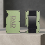 A split view of a slim metal wallet shown from the front and back on a white background. The front side is light olive green with dark gray text reading 'DAD OUR ROCK OUR STRENGTH.