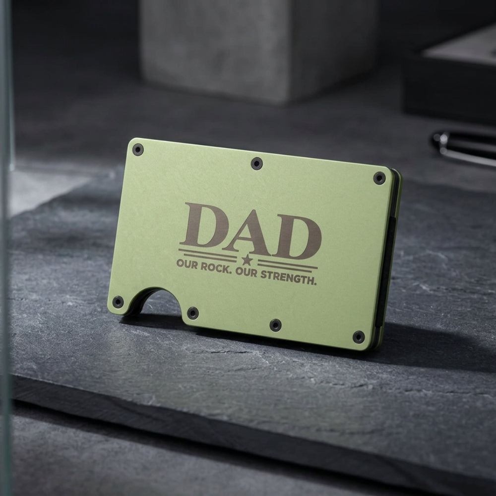 A slim, rectangular metal wallet in a muted sage green color, featuring the text 'DAD OUR ROCK. OUR STRENGTH.