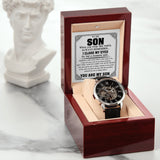 A skeleton wristwatch with a black strap is displayed inside an open reddish-brown presentation box on a light marble surface. The inside lid has a Father's Day card, and a white classical bust sculpture sits blurred in the background.