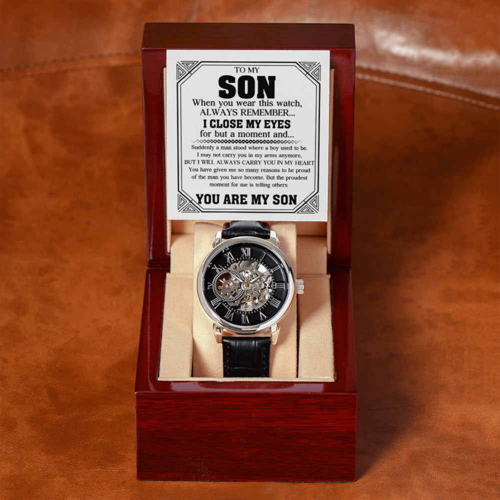A skeleton wristwatch with a black dial and black strap displayed inside an open dark wood presentation box. A printed sentimental message card reading 'TO MY SON' is placed above the watch on a brown leather surface.