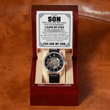 A skeleton wristwatch with a black dial and black strap displayed inside an open dark wood presentation box. A printed sentimental message card reading 'TO MY SON' is placed above the watch on a brown leather surface.