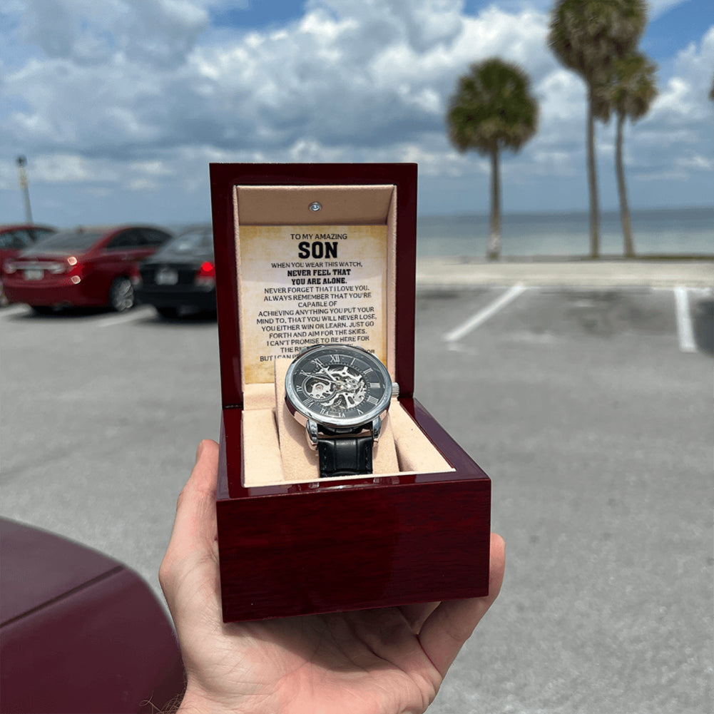 Hand holding an open mahogany-style gift box containing an automatic skeleton watch and To My Amazing Son message card outdoors.