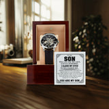 A skeleton-style wristwatch with a black dial and dark strap displayed in an open wooden presentation box, alongside a printed message card addressed 'To My Son.' The scene is set on a wooden table with a softly blurred indoor background.