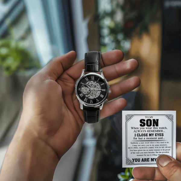 A skeleton-style wristwatch with a black dial and black leather strap is held in a person's hand, while a small memorial card addressed 'TO MY SON' is shown in the other hand. The background is a softly blurred real-world setting.