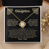 A gold necklace with a heart-shaped pendant is displayed on a black gift box insert addressed 'TO MY Daughter' with a sentimental message. The necklace is centered on the black card, with a black box lid behind it and dried flowers on a light wood surface around the box.