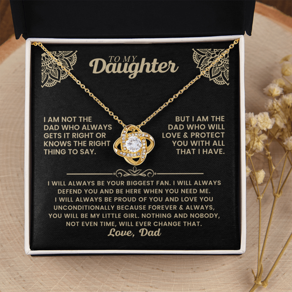 A gold necklace with a heart-shaped pendant is displayed on a black gift box insert addressed 'TO MY Daughter' with a sentimental message. The necklace is centered on the black card, with a black box lid behind it and dried flowers on a light wood surface around the box.