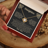 A gold necklace with a heart-shaped crystal pendant is displayed inside a red gift box on a black message card addressed to 'My Daughter.' A hand with red nail polish holds the box, and dried flowers sit beside it on a wood slice background.
