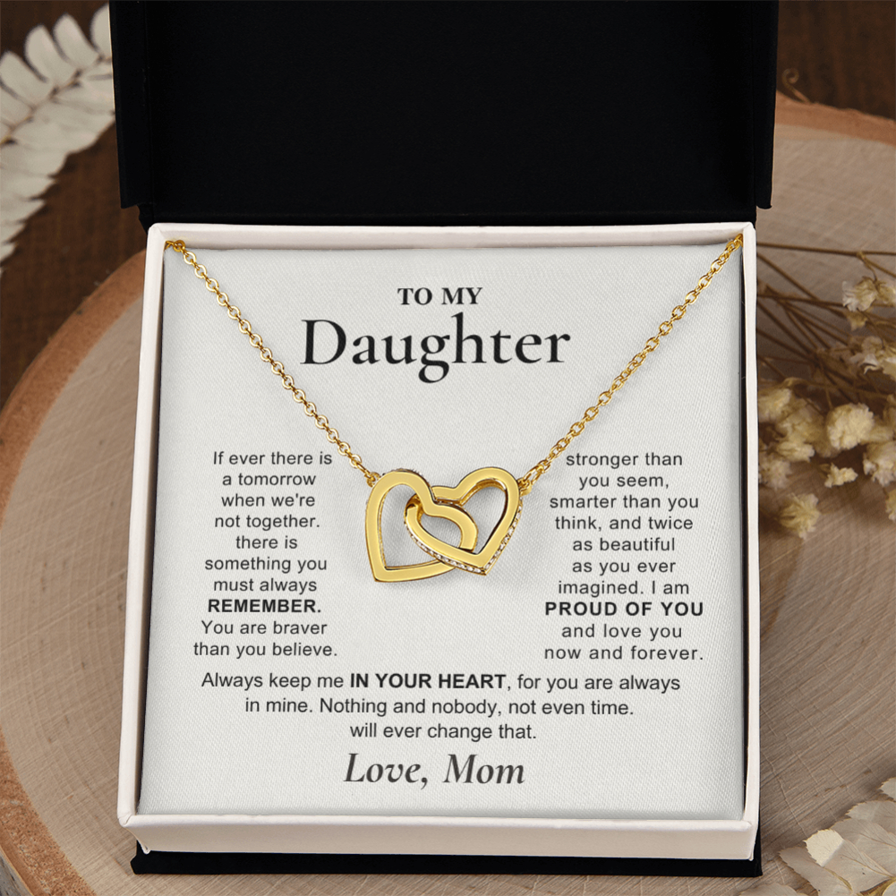 A gold double-heart pendant necklace presented in an open black gift box with a white message card addressed 'TO MY Daughter.' The necklace is centered on the card, and the box is photographed against a softly styled neutral background with dried flowers.