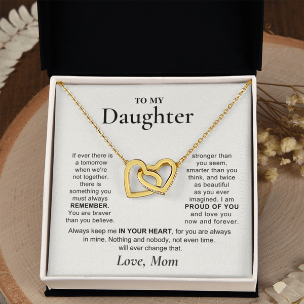 A gold double-heart pendant necklace presented in an open black gift box with a white message card addressed 'TO MY Daughter.' The necklace is centered on the card, and the box is photographed against a softly styled neutral background with dried flowers.