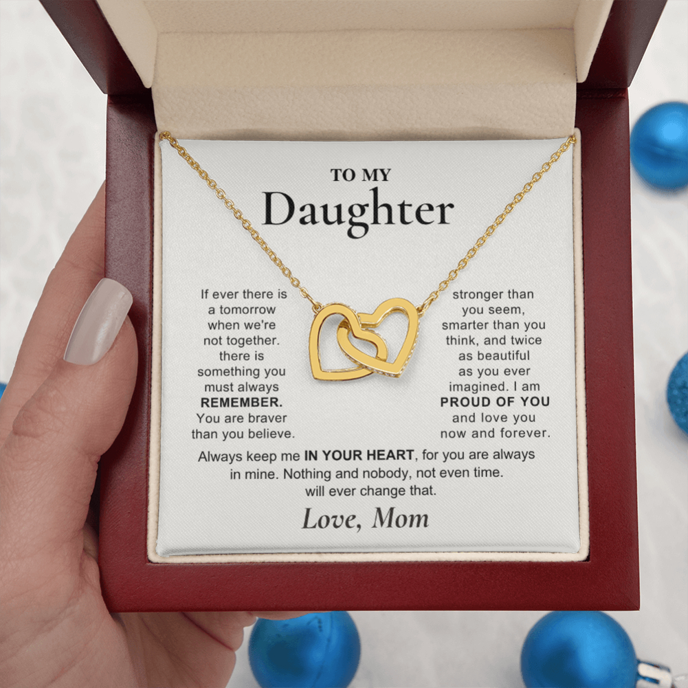 A gold heart-link necklace displayed on a printed message card inside a red gift box, held by a hand. The card is addressed 'TO MY Daughter' with a sentimental message, and the necklace features two interlocking gold hearts.