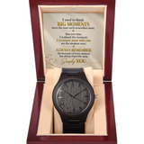 A black wooden wristwatch with a textured wood grain dial and black strap, presented inside an open dark red wooden gift box with a beige cushion. The inside lid features a sentimental message printed in gold and black text.