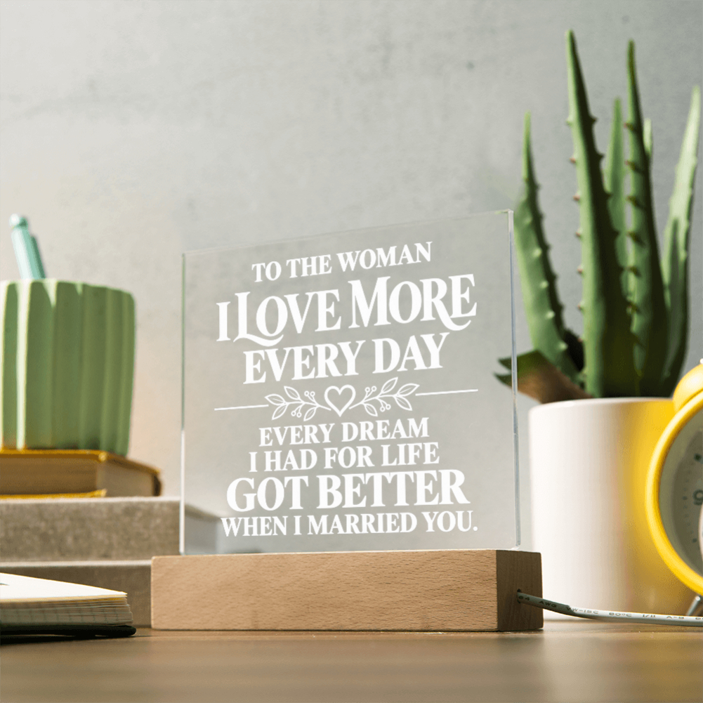 A personalized acrylic plaque on a wooden base, engraved with the sentiment 'TO THE WOMAN I LOVE MORE EVERY DAY EVERY DREAM I HAD FOR LIFE GOT BETTER WHEN I MARRIED YOU.' displayed on a desk with a potted aloe and other home decor items in the background.
