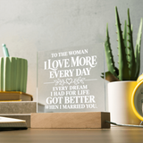 A personalized acrylic plaque on a wooden base, engraved with the sentiment 'TO THE WOMAN I LOVE MORE EVERY DAY EVERY DREAM I HAD FOR LIFE GOT BETTER WHEN I MARRIED YOU.' displayed on a desk with a potted aloe and other home decor items in the background.