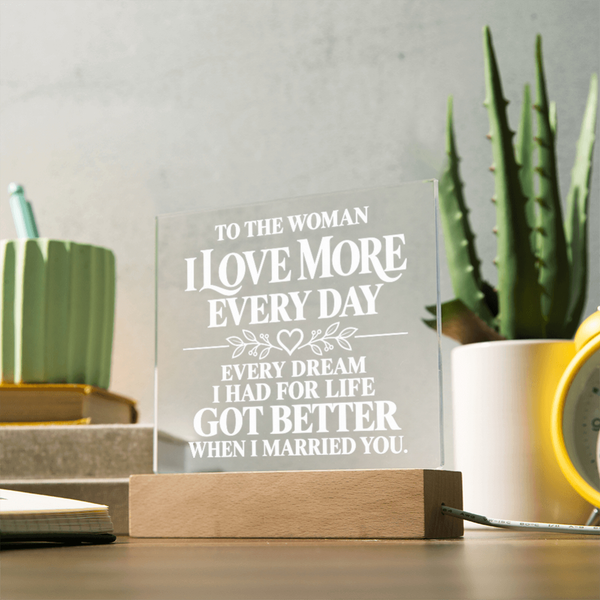 A personalized acrylic plaque on a wooden base, engraved with the sentiment 'TO THE WOMAN I LOVE MORE EVERY DAY EVERY DREAM I HAD FOR LIFE GOT BETTER WHEN I MARRIED YOU.' displayed on a desk with a potted aloe and other home decor items in the background.