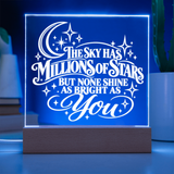 A glowing acrylic LED night light on a wooden base, displaying the quote 'THE SKY HAS MILLIONS OF STARS BUT NONE SHINE AS BRIGHT AS You' in white against a blue illuminated background.