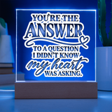 A glowing acrylic LED sign on a wooden base with the phrase “YOU’RE THE ANSWER TO A QUESTION I DIDN’T KNOW MY HEART WAS ASKING.” The sign is lit in blue and white, shown on a tabletop with blurred plants in the background.