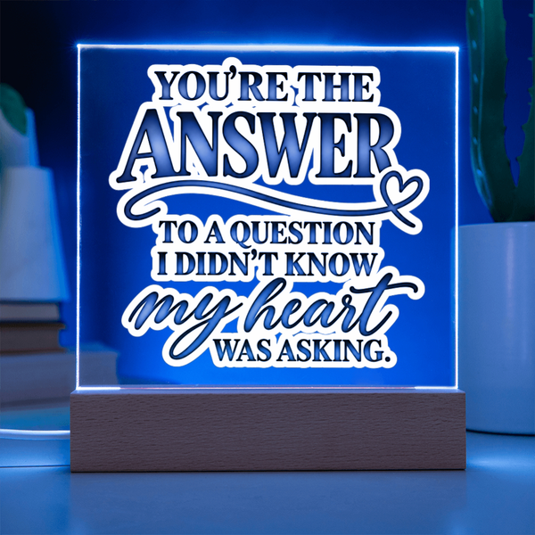 A glowing acrylic LED sign on a wooden base with the phrase “YOU’RE THE ANSWER TO A QUESTION I DIDN’T KNOW MY HEART WAS ASKING.” The sign is lit in blue and white, shown on a tabletop with blurred plants in the background.