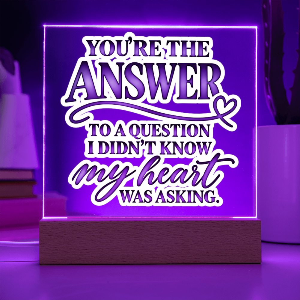 A rectangular LED acrylic sign on a wooden base glowing with purple light, featuring the quote 'YOU'RE THE ANSWER TO A QUESTION I DIDN'T KNOW my heart WAS ASKING.' in white and purple lettering.