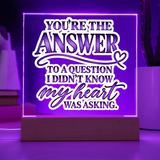 A rectangular LED acrylic sign on a wooden base glowing with purple light, featuring the quote 'YOU'RE THE ANSWER TO A QUESTION I DIDN'T KNOW my heart WAS ASKING.' in white and purple lettering.