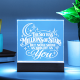 A square LED acrylic night light with a wooden base, glowing with a blue light and featuring the quote 'THE SKY HAS MILLIONS OF STARS BUT NONE SHINE AS BRIGHT AS You' in white script. It is shown on a tabletop next to a potted plant and books, with a dark blue background.