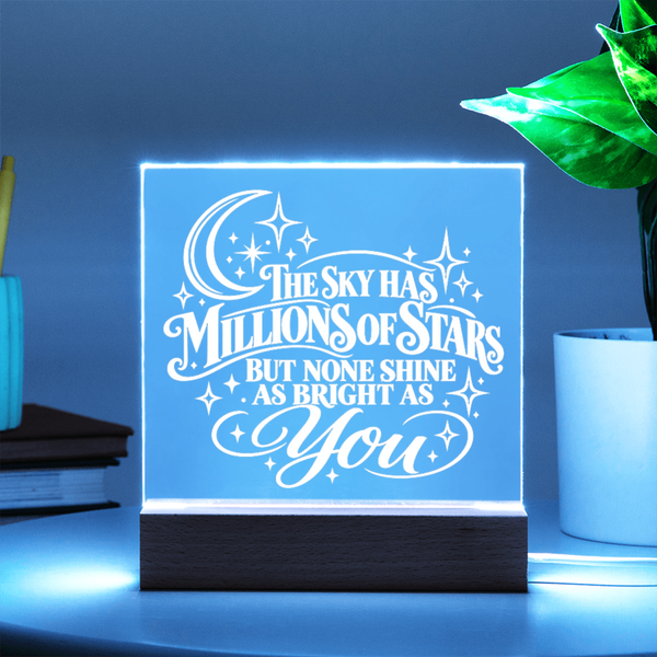 A square LED acrylic night light with a wooden base, glowing with a blue light and featuring the quote 'THE SKY HAS MILLIONS OF STARS BUT NONE SHINE AS BRIGHT AS You' in white script. It is shown on a tabletop next to a potted plant and books, with a dark blue background.
