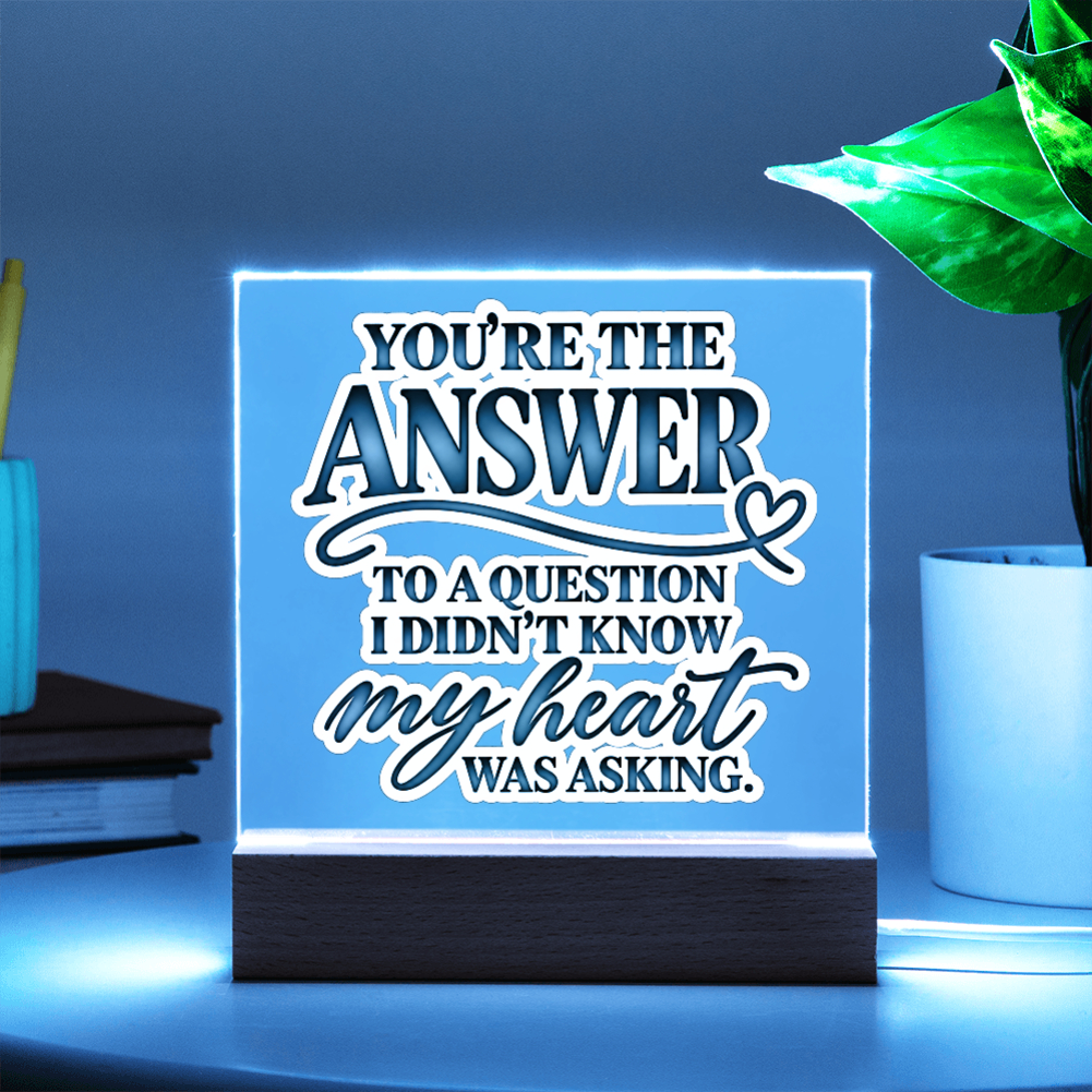 A square acrylic LED sign on a dark wooden base, glowing with a blue light. The sign features white and dark blue text that reads, 'YOU'RE THE ANSWER TO A QUESTION I DIDN'T KNOW my heart WAS ASKING.
