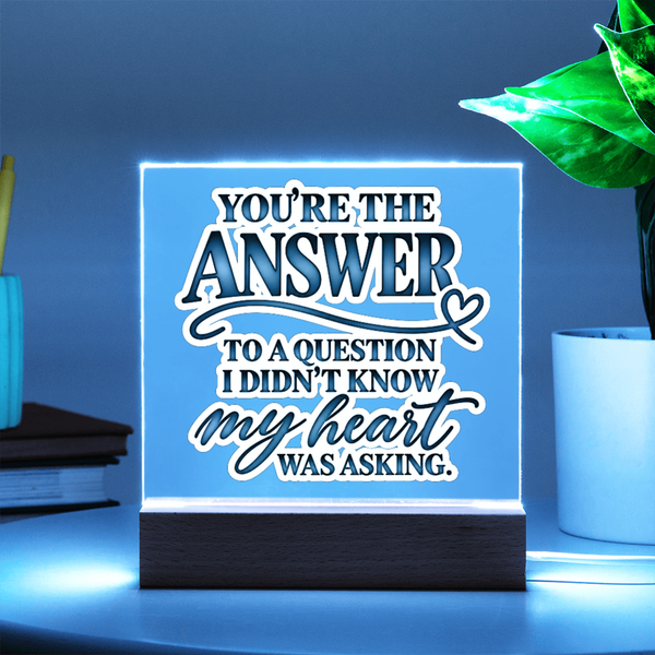 A square acrylic LED sign on a dark wooden base, glowing with a blue light. The sign features white and dark blue text that reads, 'YOU'RE THE ANSWER TO A QUESTION I DIDN'T KNOW my heart WAS ASKING.