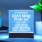 A glowing acrylic LED sign on a wooden base with white text that reads, 'TO THE WOMAN I LOVE MORE EVERY DAY EVERY DREAM I HAD FOR LIFE GOT BETTER WHEN I MARRIED YOU.' The sign is shown lit in a blue-toned room setting on a tabletop, with a plant and books in the background.