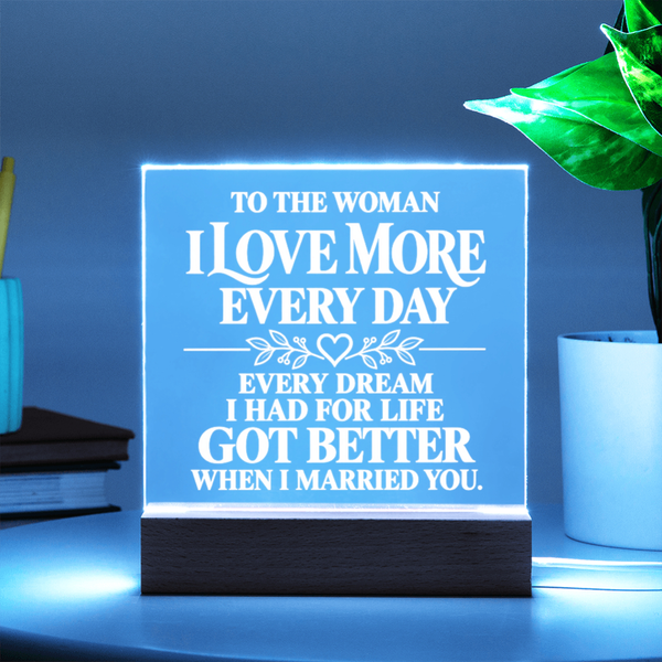 A glowing acrylic LED sign on a wooden base with white text that reads, 'TO THE WOMAN I LOVE MORE EVERY DAY EVERY DREAM I HAD FOR LIFE GOT BETTER WHEN I MARRIED YOU.' The sign is shown lit in a blue-toned room setting on a tabletop, with a plant and books in the background.