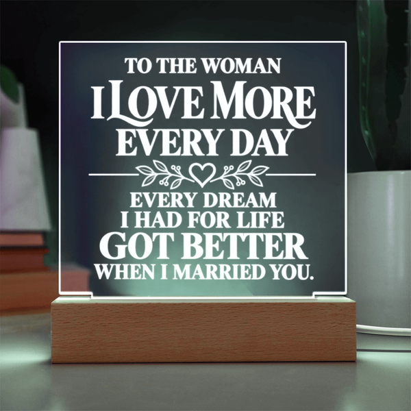 A rectangular acrylic LED sign with white text reading 'TO THE WOMAN I LOVE MORE EVERY DAY EVERY DREAM I HAD FOR LIFE GOT BETTER WHEN I MARRIED YOU.' displayed on a wooden base in a home setting.
