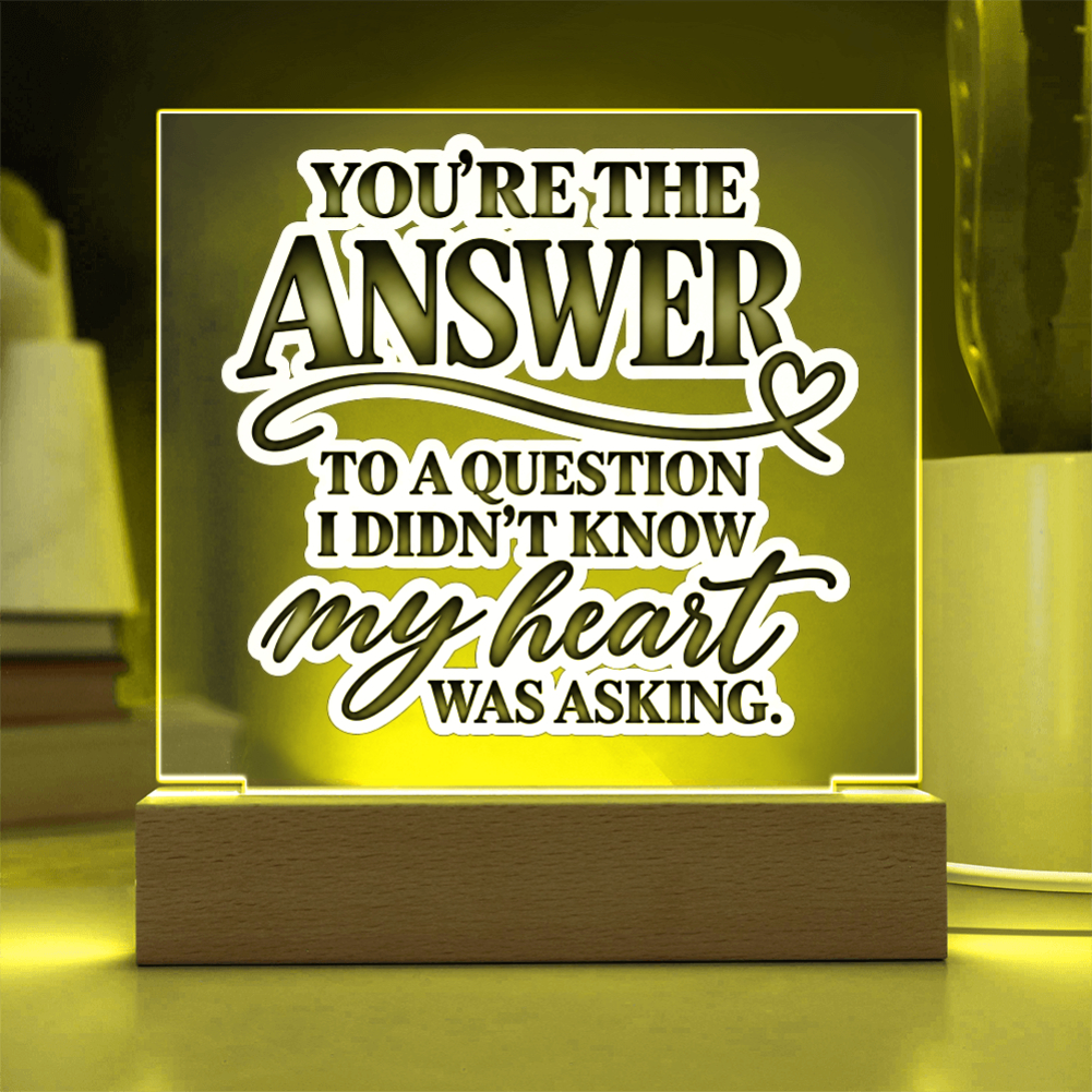 A square acrylic LED message lamp with a wooden base, glowing with a warm yellow light. The clear acrylic panel features the printed quote, 'YOU'RE THE ANSWER TO A QUESTION I DIDN'T KNOW my heart WAS ASKING.