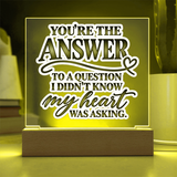 A square acrylic LED message lamp with a wooden base, glowing with a warm yellow light. The clear acrylic panel features the printed quote, 'YOU'RE THE ANSWER TO A QUESTION I DIDN'T KNOW my heart WAS ASKING.