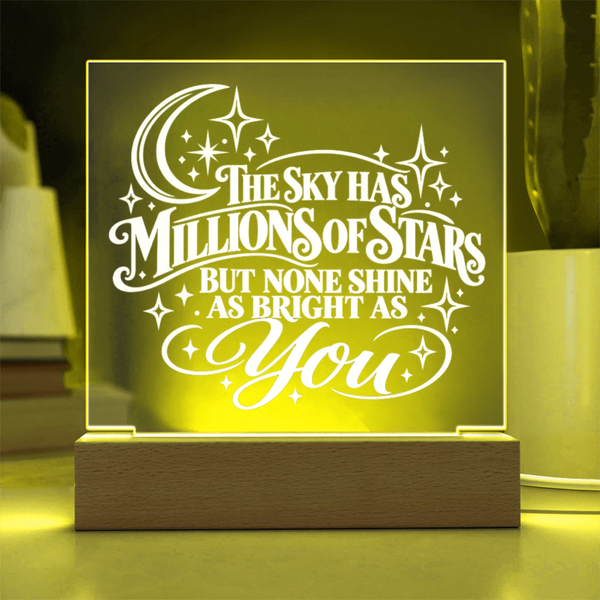 A glowing acrylic LED night light with a wooden base, featuring the quote 'THE SKY HAS MILLIONS OF STARS BUT NONE SHINE AS BRIGHT AS You' in white script and star motifs, set against a warm yellow light.