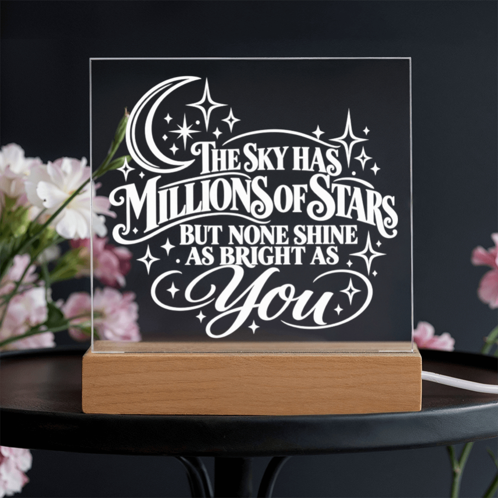 A wooden LED night light with a clear acrylic panel displaying the quote 'The Sky Has Millions of Stars But None Shine As Bright As You' in white lettering, set on a black table with blurred pink flowers in the background.