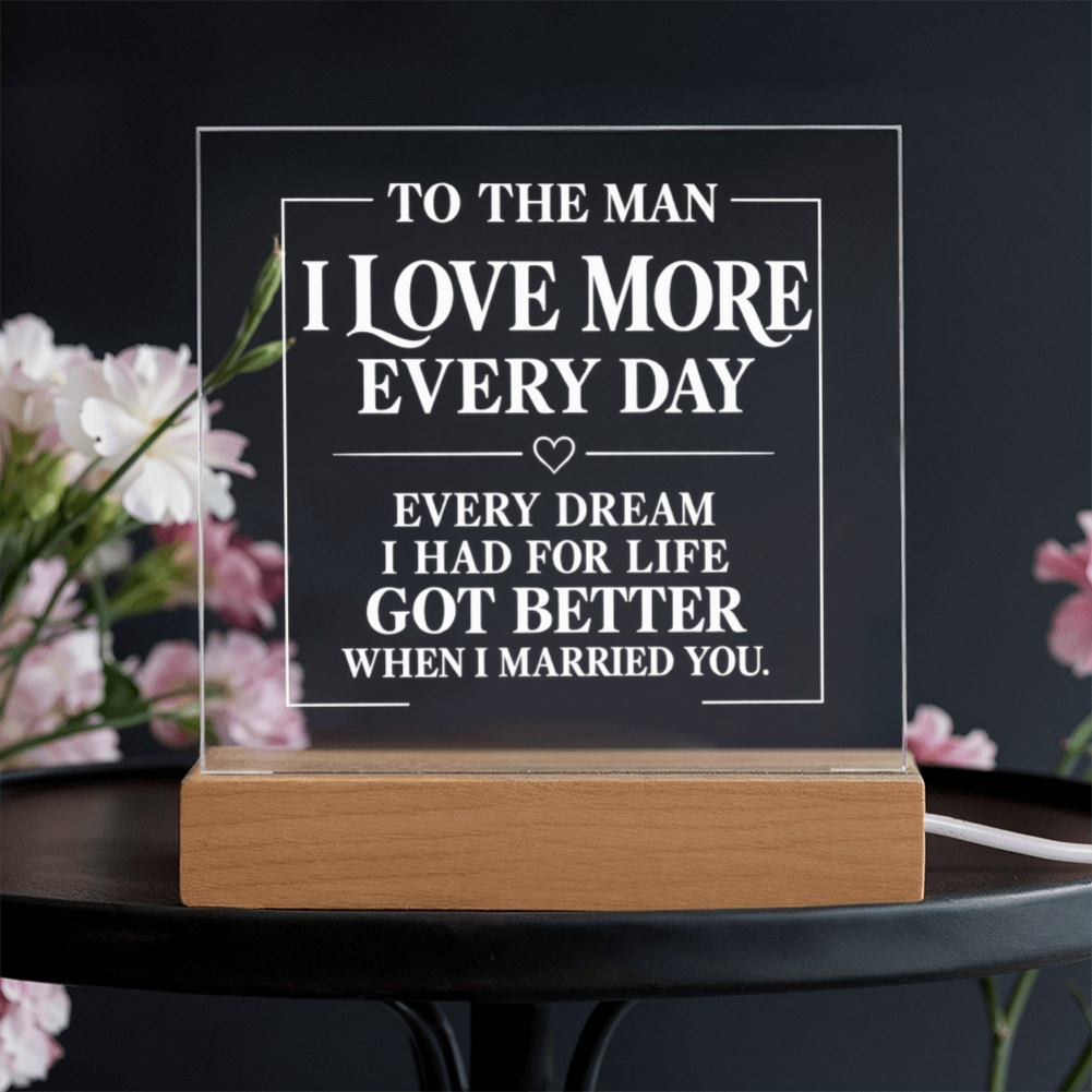A rectangular acrylic LED light box with engraved white text reading 'TO THE MAN I LOVE MORE EVERY DAY' and a romantic message below, resting on a light wood base on a dark table with blurred pink flowers in the background.