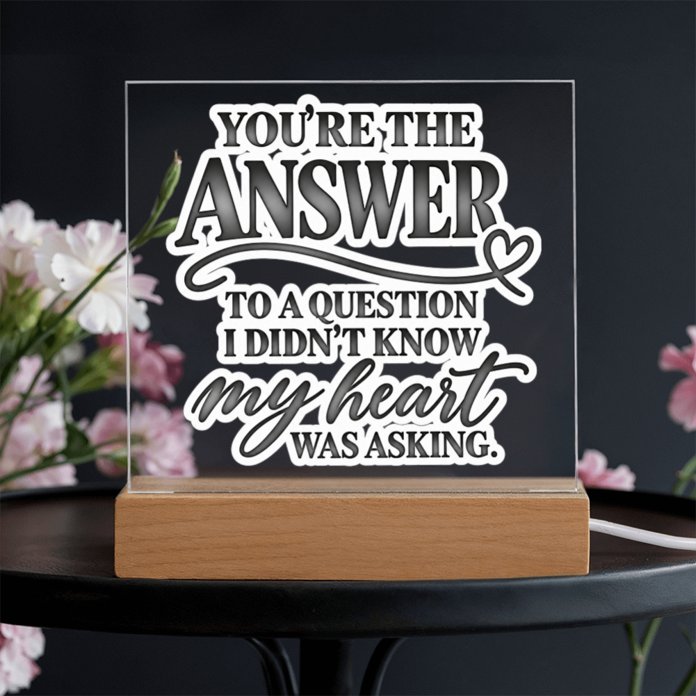 A rectangular acrylic LED light sign with a wooden base, featuring the quote 'YOU'RE THE ANSWER TO A QUESTION I DIDN'T KNOW MY HEART WAS ASKING.' in white and gray lettering, displayed on a dark table with blurred pink flowers in the background.