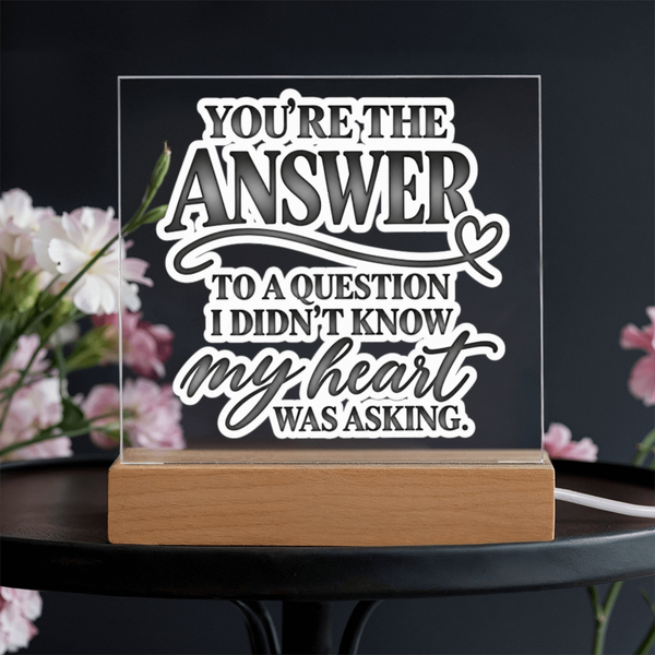 A rectangular acrylic LED light sign with a wooden base, featuring the quote 'YOU'RE THE ANSWER TO A QUESTION I DIDN'T KNOW MY HEART WAS ASKING.' in white and gray lettering, displayed on a dark table with blurred pink flowers in the background.