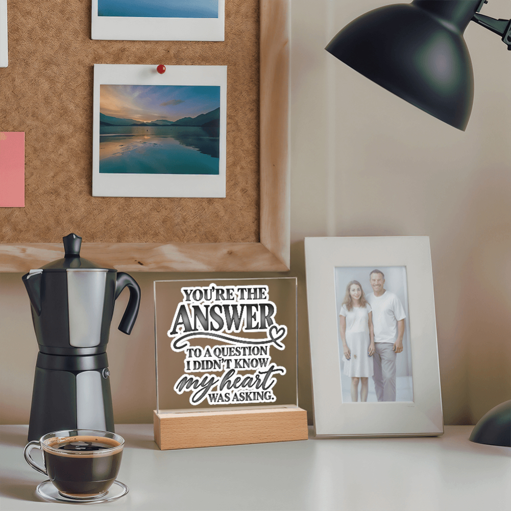 A personalized acrylic sign on a wooden base that reads 'YOU'RE THE ANSWER TO A QUESTION I DIDN'T KNOW my heart WAS ASKING.' styled on a desk next to a moka pot, coffee cup, and a framed photo of a couple, with a cork board and lamp in the background.