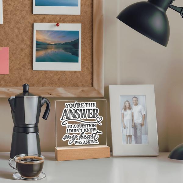 A personalized acrylic sign on a wooden base that reads 'YOU'RE THE ANSWER TO A QUESTION I DIDN'T KNOW my heart WAS ASKING.' styled on a desk next to a moka pot, coffee cup, and a framed photo of a couple, with a cork board and lamp in the background.