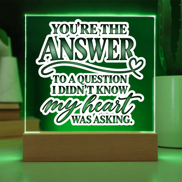 A square acrylic LED sign on a wooden base glowing green with white text that reads, 'YOU'RE THE ANSWER TO A QUESTION I DIDN'T KNOW MY HEART WAS ASKING.' The sign is photographed in a styled indoor setting with soft green lighting and blurred background objects.