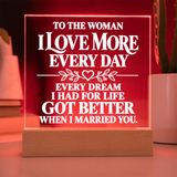 A rectangular acrylic LED sign on a wooden base with white text reading, 'TO THE WOMAN I LOVE MORE EVERY DAY EVERY DREAM I HAD FOR LIFE GOT BETTER WHEN I MARRIED YOU,' shown glowing red in a home setting.